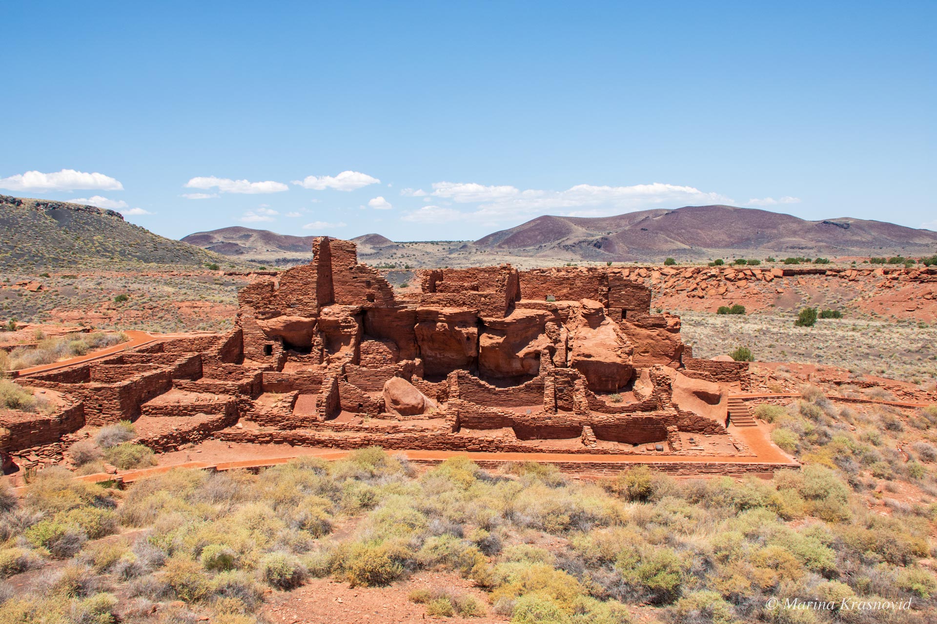 Ancient red sandstone pueblo ruins at Wupatki National Monument, surrounded by desert scrub and distant volcanic peaks under a bright Arizona sky.