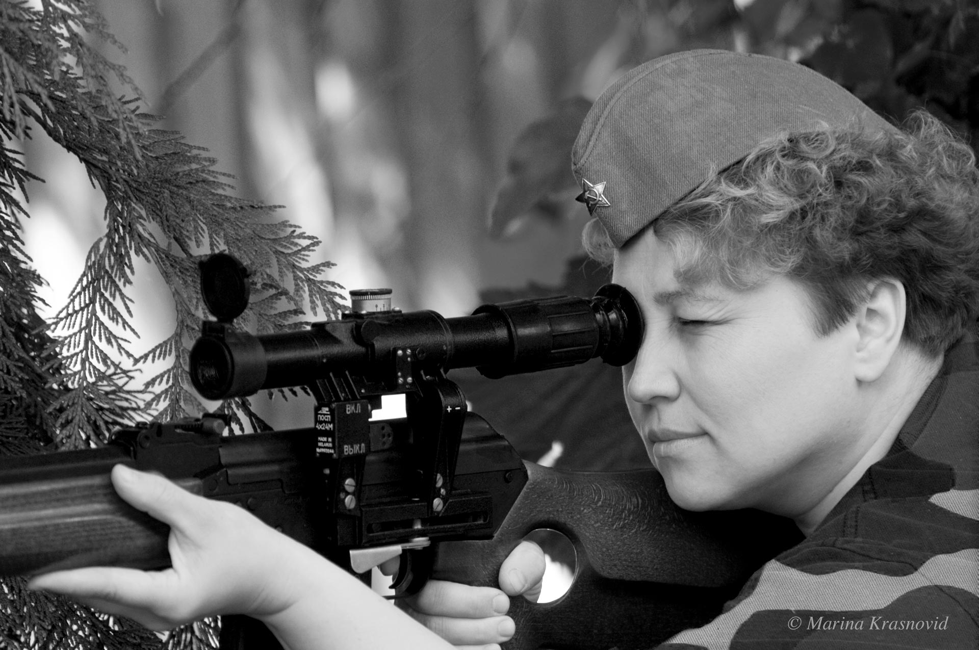 Woman in a Soviet-era uniform aiming through a sniper scope in a black and white portrait, symbolizing focus and strength. Photographed by Marina Krasnovid.