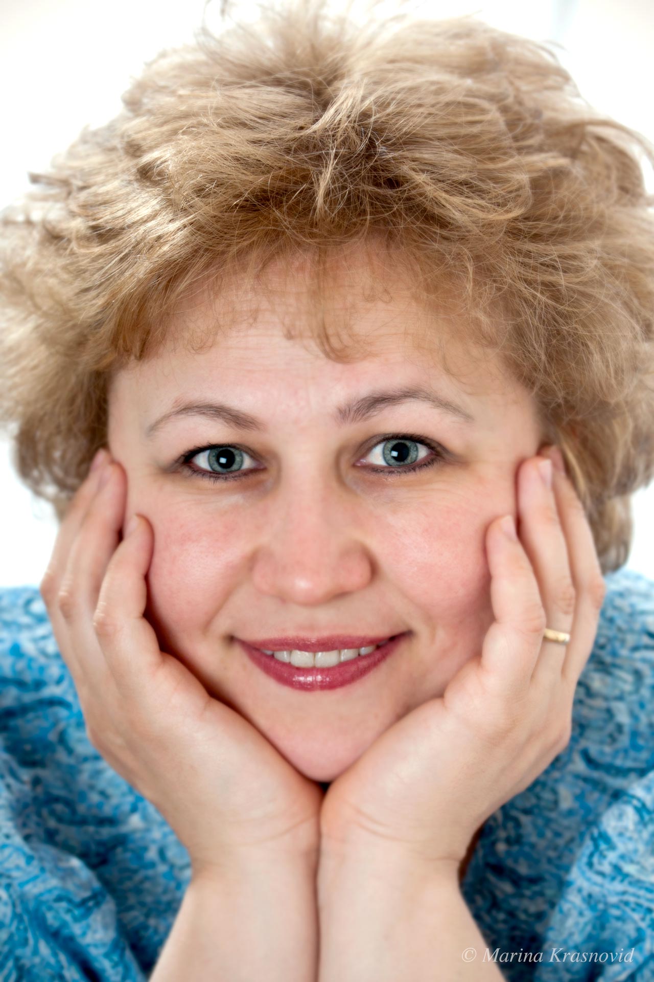 Through Kind Eyes Close-up portrait of a woman smiling softly with her hands resting on her cheeks, wearing a blue patterned blouse. Photographed by Marina Krasnovid.