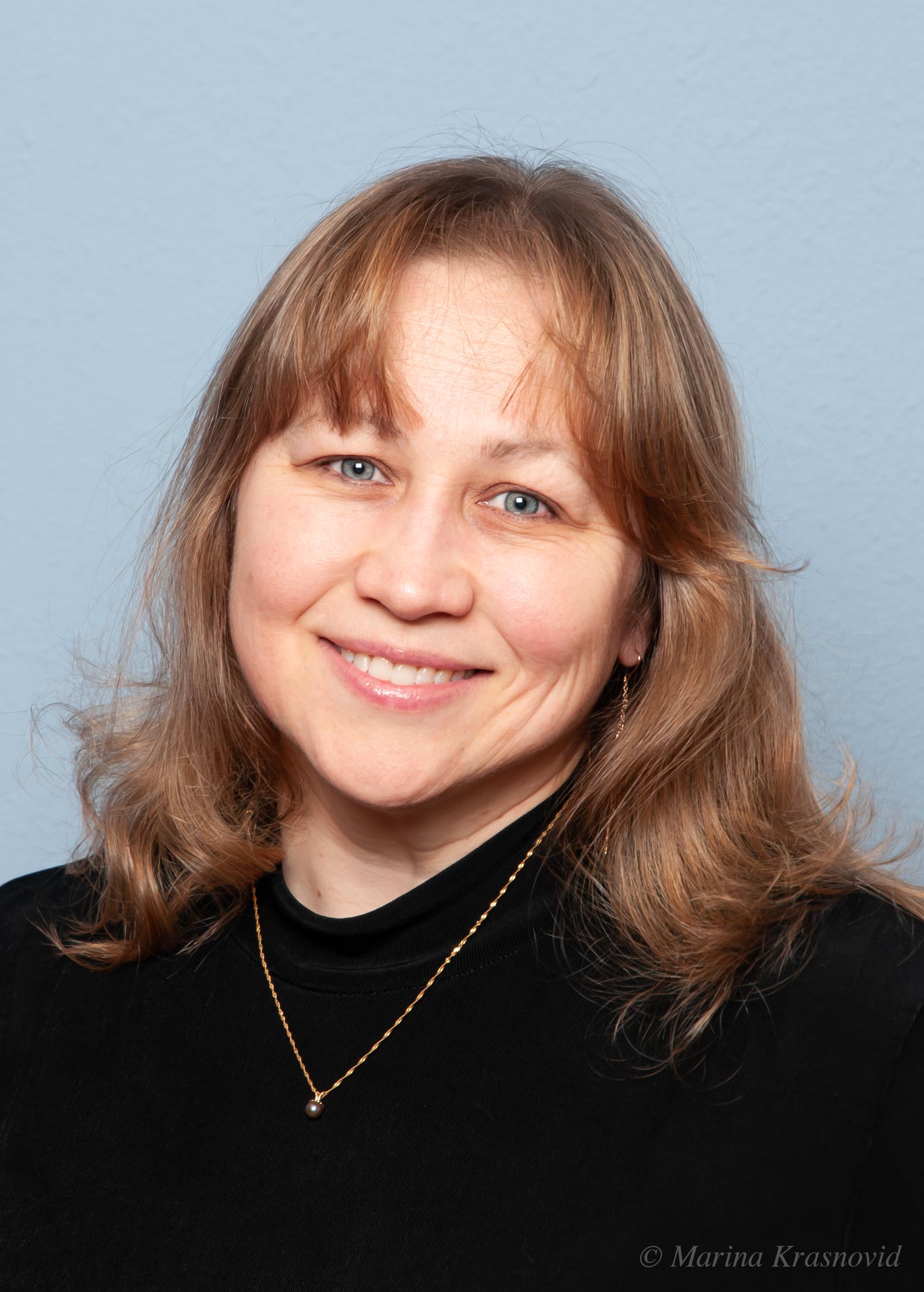 A Moment of Lightness Smiling woman with light brown hair and blue eyes wearing a black top and gold necklace, photographed against a soft blue background. Photographed by Marina Krasnovid.