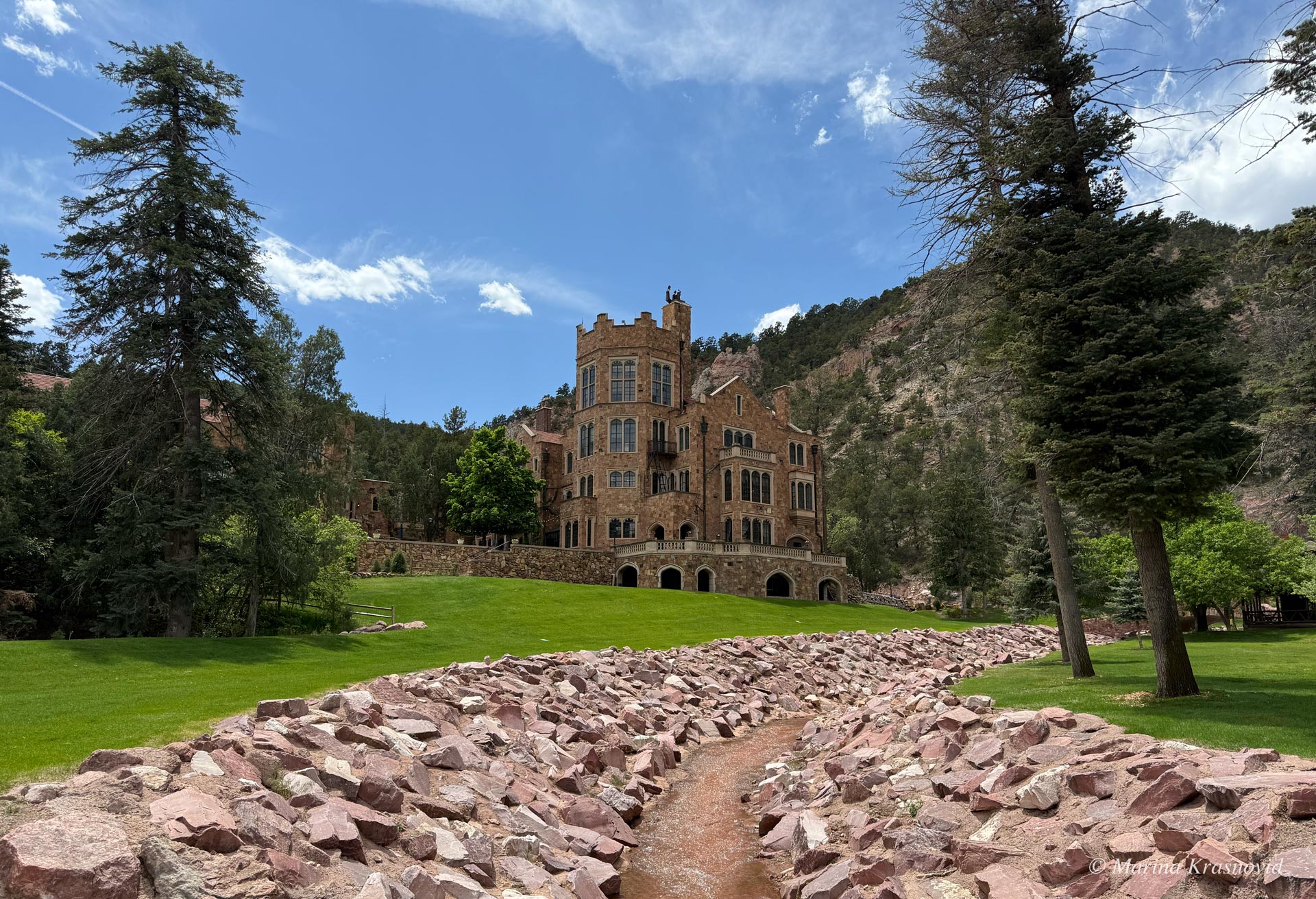 Historic Glen Eyrie Castle in Colorado Springs, Colorado, surrounded by pine-covered mountains and green lawns with a red rock creek in the foreground | photo by Marina Krasnovid
