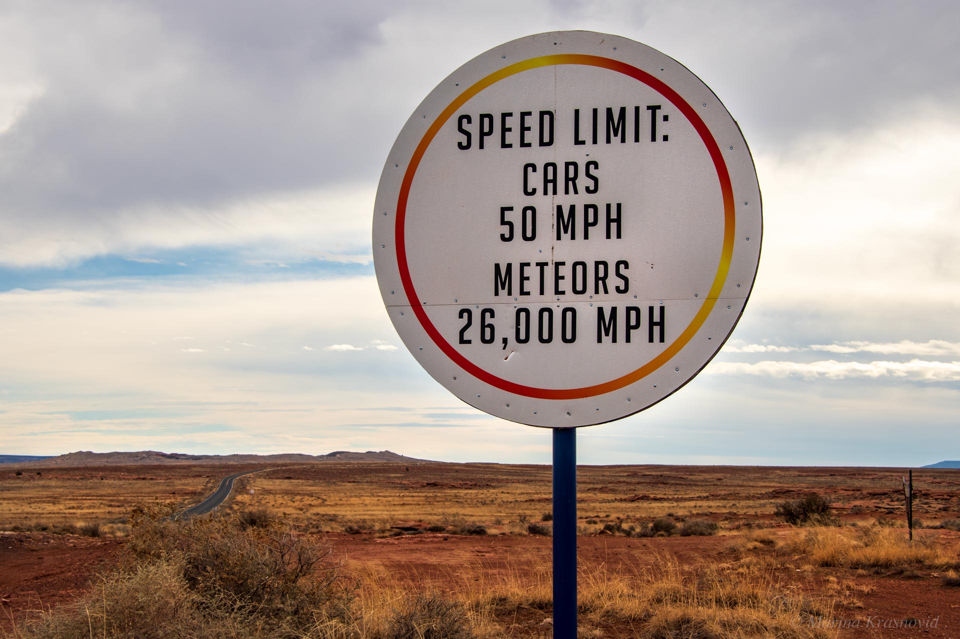 Roadside sign near Meteor Crater in Arizona reading “Speed Limit: Cars 50 MPH, Meteors 26,000 MPH,” with open desert landscape in the background.